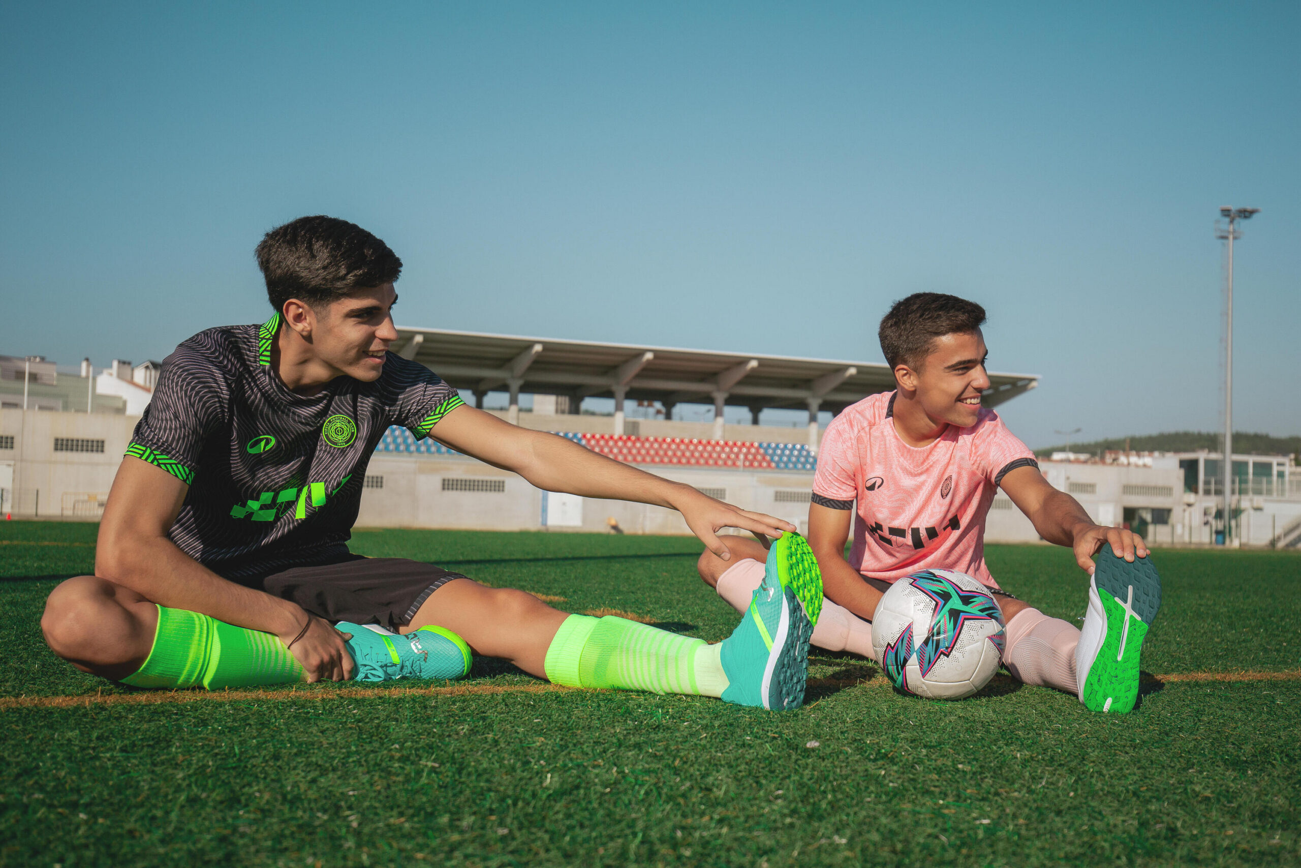 Two male footballers stretching in custom Decathlon football kit; one green and black, one pink and grey.