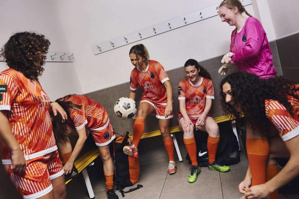 Female football players in the changing rooms wearing orange and white matching patterned shorts and shirts, with orange socks.