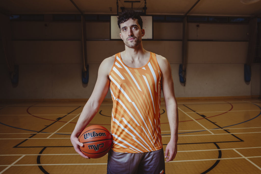 Basketball player in custom orange vest and grey shorts in an indoor court, holding a basketball